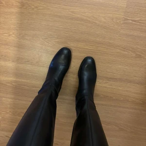 Svarta skinnstövlar från Pace, stl 39 - Klassiska svarta stövlar i skinn från Pace med låg klack och rund tå. Snygg, stilren design som går upp till knäet och passar perfekt till både jeans och kjol. Sula i syntetmaterial och insida i skinn. Tidlösa boots för dig som gillar minimalistisk stil.