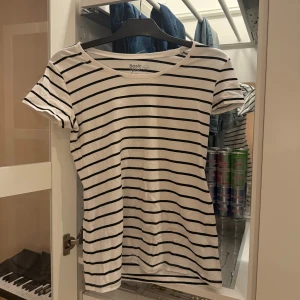 Randig vit t-shirt - Vit t-shirt med svarta horisontella ränder från Basic Apparel. Klassisk rund halsringning och korta ärmar. Skön och lätt i materialet, perfekt till jeans eller shorts. Enkel och tidlös design som funkar året runt.