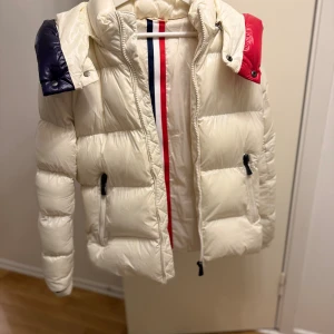 Vit pufferjacka med färgdetaljer - Vit puffjacka i fint skick. Jackan har blå och röda detaljer som ger en stilren och modern look. Den är varm, bekväm och passar bra till både vardag och fritid. 	•	Dragkedja framtill 	•	Två fickor 	•	Luvan har blå och röda detaljer  En praktisk och snygg jacka för kalla dagar