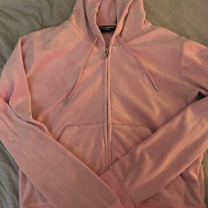 Rosa velour hoodie från Juicy Couture - Supermjuk rosa hoodie från Juicy Couture i klassisk velour med dragkedja och huva. Snygga detaljer som snörning i halsen, fickor framtill och ett J-format dragkedjesmycke. Perfekt för dig som vill ha en ikonisk och bekväm look.