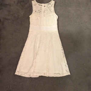 Vit spetsklänning med ärmlös design - Superfin vit klänning i spets med ärmlös modell och rund halsringning. Klänningen har markerad midja och lätt utställd kjol.