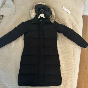 Svart lång dunjacka från Everest - Säljer en svart lång dunjacka från Everest med mysig fuskpäls på huvan. Jackan har dragkedja och knappar framtill, två fickor och quiltad design. Perfekt för kalla dagar när du vill hålla dig varm och ändå se snygg ut. Använd bara 2-3 gånger. Pris i butik 3000kr och uppåt