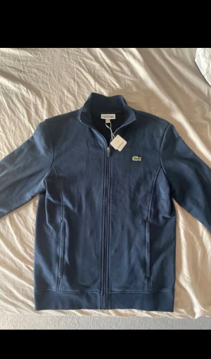 Marinblå zipjacka från Lacoste Sport - Helt ny med ordinarie pris (1600)Snygg marinblå zipjacka från Lacoste Sport med klassisk krokodillogga på bröstet. Jackan har hög krage, hel dragkedja och två fickor framtill. Tillverkad i mjuk bomullsmix för en bekväm känsla. Perfekt för en sportig och clean look.