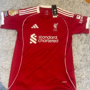 Liverpool röd fotbollströja Adidas - Liverpool FC matchtröja från Adidas i röd färg med vita detaljer och tryck. Kortärmad med klassiska tre ränder på axlarna, Premier League-märke på ärmen och 'ISAK 9' på ryggen. Materialet är lätt och andas, Aeroready-teknologi. Snyggt LFC-emblem på bröstet.