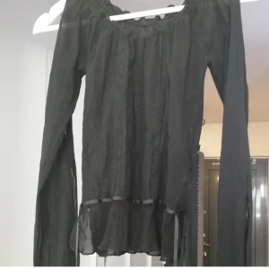 Svart transparent blus med volang - Säljer en svart transparent blus med långa ärmar och snygga slitsar längs armarna. Blusen har volangdetalj nedtill och spets längs framstycket, snörren vid midjan. Köpt i början på 2000-talet, men inte använts.