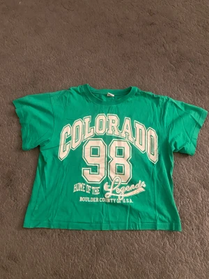 Grön Colorado t-shirt från Lindex - Cool grön t-shirt från Lindex med stort vitt tryck 'COLORADO 98 Home of the Legends Boulder County of U.S.A.' på framsidan. Klassisk rund hals och korta ärmar, perfekt för dig som gillar sportig och avslappnad stil. Materialet är mjuk bomull.