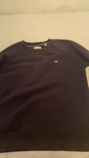 Mörkblå sweatshirt från GANT - Säljer en stilren mörkblå sweatshirt från GANT med rund hals och diskret logga broderad på bröstet. Tröjan har långa ärmar och ribbade muddar vid hals, ärmslut och nederkant. Perfekt för dig som gillar klassisk och enkel design.Villig att sänka pris!