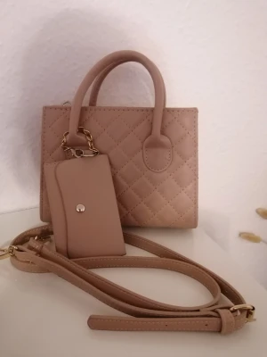 Beige quiltad handväska med plånbok - Snygg beige handväska med quiltad design och matchande plånbok. Väskan har både handtag och justerbar axelrem med guldfärgade detaljer. Materialet är skinnimitation och väskan har en stilren, rektangulär form. Perfekt för dig som gillar minimalistisk och chic stil. Den är i väldigt bra skick (har använt x antal gånger) . Men plånbokens kedja är lite rostad, men inget man märker. ☺️💞