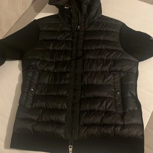 Svart Moncler pufferjacka med huva - Snygg svart pufferjacka från Moncler med huva och ribbade ärmar. Jackan har två stora fickor framtill med knappar, dragkedja i mitten och en extra ficka med dragkedja på ärmen. Klassisk Moncler-logga på ärmen och coolt innerfoder med serietidningsmotiv.
