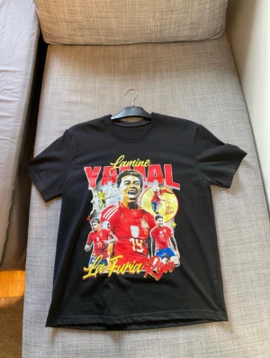 Svart Lamine Yamal t-shirt med tryck - Cool svart t-shirt med färgstarkt tryck av Lamine Yamal och texten 'La Furia Roja' i gult och rött. Motiv med flera fotbollsbilder och stora röda bokstäver. Rund hals och korta ärmar, perfekt för dig som älskar fotboll och vill sticka ut.