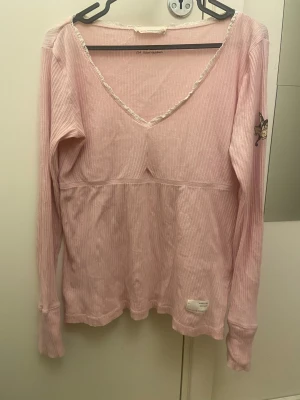 Rosa ribbad Odd Molly långärmad tröja - Supersöt ljusrosa långärmad tröja från Odd Molly med ribbad struktur och v-ringad hals med spetskant. På ena ärmen finns en snygg fjärilsdetalj med Odd Molly-logga. Tröjan är i mjuk bomull och har normal passform, perfekt för dig som gillar detaljer. Jag skulle såga att den är Storlek S men kan även passa i M