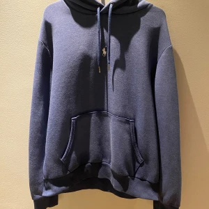 Blå hoodie från Polo Ralph Lauren - Mysig blå hoodie från Polo Ralph Lauren med klassisk huva, dragsnören med metalländar och broderad logga på snöret. Tröjan har känguruficka framtill och ribbade muddar vid ärmslut och nederkant. Perfekt för chill dagar. Jag är 180 lång och den sitter fint liten fläck finns 
