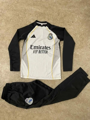 Real Madrid långärmad fotbollströja Adidas - Säljer en långärmad Real Madrid fotbollströja från Adidas i svart och vitt med gula detaljer. Tröjan har klubbmärke och sponsortryck på bröstet, samt dragkedja vid halsen. Materialet är lätt och andas, perfekt för träning eller match.
