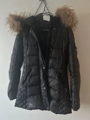 Svart Rockandblue pufferjacka med fuskpäls - Svart pufferjacka från Rockandblue med quiltad design, dragkedjor på fickor och ärmslut samt badge på ärmen. Jackan har en stor huva med brun fuskpäls och snygga detaljer i metall. Perfekt för kalla dagar och riktigt stilren look.