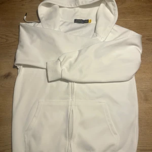 Vit hoodie från Polo Ralph Lauren stl m - Vit hoodie från Polo Ralph Lauren med dragkedja och klassisk huva. Tröjan har ribbade muddar, fickor framtill och den ikoniska Polo-loggan broderad på bröstet. Tillverkad i mjuk bomull för skön komfort. Perfekt för chill dagar och streetwear-stil.