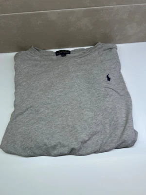 Grå långärmad tröja Polo Ralph Lauren - Snygg grå långärmad tröja från Polo Ralph Lauren i storlek BARN XL. Tröjan har en klassisk rund hals och är tillverkad i mjuk bomull. På bröstet finns den ikoniska broderade Polo-loggan i mörkblått. Perfekt för en chill och stilren look.