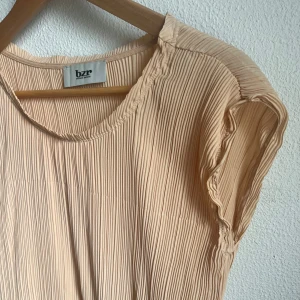 Plisserad klänning dansk design - Superfin plisserad klänning från danska Bruuns Bazar, färg i en varm beige/ljust aprikosrosa nyans. Testa att styla t ex med eget skärp eller smycken som kontrast (bälte på bilden ingår inte i köpet). Klänningen har en löst sittande resår i midjan. Materialet är lätt och ger ett snyggt fall, insydd beige underkjol. Stilren klänning i bra kvalitet med många användningsområden. 