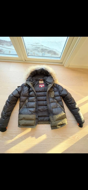 Parajumpers dunjacka med päls - Snygg Parajumpers dunjacka i svart och olivgrön med fluffig päls på huvan. Jackan har flera fickor med dragkedja, ribbade muddar och coola detaljer som gul rem vid kragen. Perfekt för kalla dagar och riktigt stilren look. Storlek 14 år 