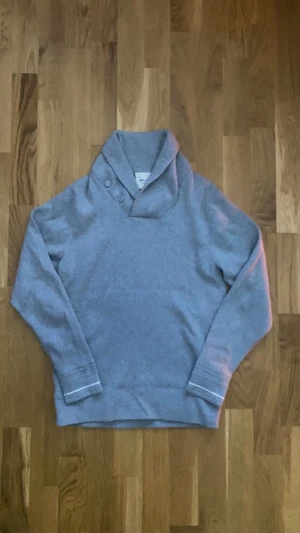 Grå stickad tröja NN07 med krage - Säljer en grå stickad tröja från NN07 i storlek S. Tröjan har en snygg omlottkrage med två knappar och är tillverkad i mjuk bomull och kashmir. Långärmad modell med diskreta ribbade muddar och stilren look. Perfekt för dig som gillar minimalistisk stil. 