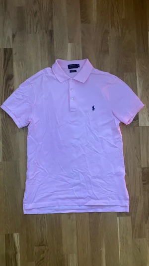 Rosa pikétröja Polo Ralph Lauren  - Snygg rosa pikétröja från Polo Ralph Lauren i slim fit med stretch mesh-material. Klassisk krage, korta ärmar och tre knappar framtill. Ikonisk marinblå logga broderad på bröstet. Perfekt för dig som gillar stilrena och sportiga plagg. Storlek M, vid frågor/funderingar tveka inte på att skriva!