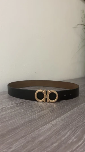 Svart skärp från Salvatore Ferragamo - Stilrent svart bälte i skinn från Salvatore Ferragamo med ikoniskt dubbel-Gancini-spänne i guld. Bältet har en klassisk, rak form och är perfekt för att lyfta vilken outfit som helst med sin lyxiga känsla och snygga detaljer.