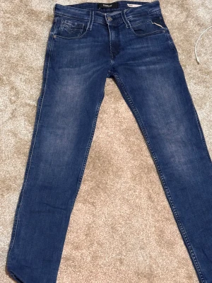 Replay Anbass slim fit jeans blå - Snygga blå Replay Anbass jeans i slim fit med klassisk femficksdesign och diskreta slitningar. Modellen har normal midja och raka ben, tillverkade i mjukt jeanstyg för skön passform. Perfekta för dig som gillar stilrena och moderna jeans. Storlek 27/30