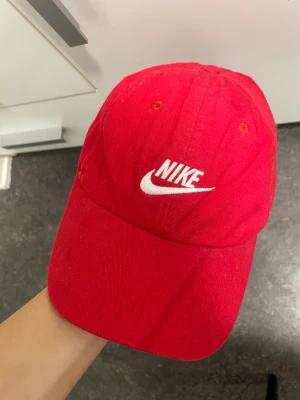 Röd Nike keps med justerbar rem - Snygg röd keps från Nike med klassisk böjd skärm och vit broderad logga framtill. Kepsen har justerbar rem med metallspänne baktill för perfekt passform. 