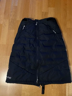 Marinblå kjol från Uhip med dragkedja - Marinblå vadderad kjol från Uhip med hel dragkedja fram och reflexdetaljer på sidorna. Kjolen har två dragkedjeförsedda fickor och är fodrad i ljusgrått. Perfekt för kalla dagar och utomhusaktiviteter.