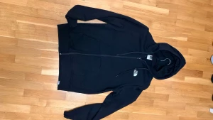 Svart hoodie från The North Face M - Svart hoodie från The North Face i storlek M med stor vit logga och bergsmotiv på ryggen samt mindre logga på bröstet. Tröjan har huva med snörning, två fickor fram och hel dragkedja. Perfekt för chill dagar och streetwear.