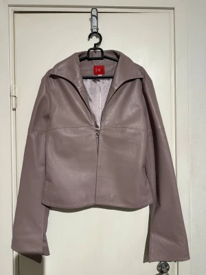 Ljuslila skinnjacka från Jag - Säljer en stilren ljuslila skinnjacka från Jag med bred krage och raka, långa ärmar. Jackan har croppad passform och stängs med hakar framtill. Snygg minimalistisk look och mjukt material med glansig insida.