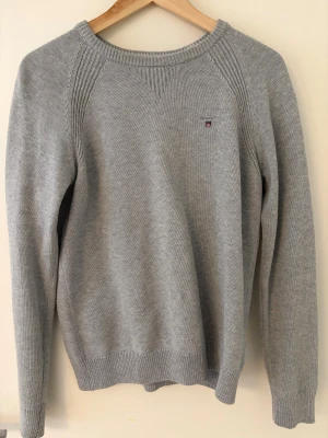 Grå stickad tröja från GANT - Snygg grå stickad tröja från GANT med diskret logga på bröstet. Tröjan har rund hals, ribbade muddar och långärmad design. Perfekt för dig som gillar klassisk och enkel stil med en touch av preppy. Materialet är mjukt och håller dig varm under kyliga dagar.