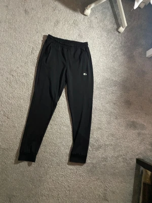 Svarta mjukisbyxor från Lacoste - Svarta mjukisbyxor från Lacoste med klassisk krokodillogga på höften. Byxorna har elastisk midja, smal passform och ribbade muddar vid bensluten. Perfekta för chill eller träning, med mjukt material och diskreta fickor. Riktigt sköna men inte helt min stil. Pris kan diskuteras!!
