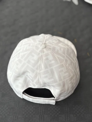 Vit keps från Fendi med FF-mönster - Snygg vit keps från Fendi med det klassiska FF-logomönstret över hela kepsen. Kepsen har böjd skärm och justerbar rem baktill. Tillverkad i slitstark bomull med exklusiv känsla, perfekt för dig som gillar designeraccessoarer.