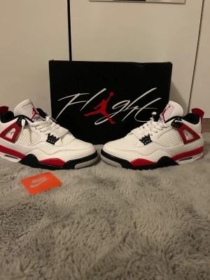 Nike Air Jordan 4 red cement  - Nike Air Jordan 4 Red cement med vit ovandel i skinn, röda och svarta detaljer samt meshpaneler på sidorna. Klassisk Jumpman-logga på plösen och Air Jordan-etikett på insidan. Snygg och ikonisk siluett med platt sula och snörning.