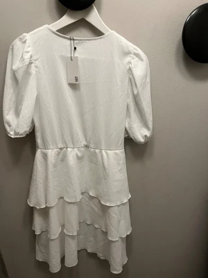 Vit volangklänning Fiora star - Supersöt vit klänning med flera volanger och puffärmar. Klänningen har rund halsringning och är tillverkad i ett lätt, strukturerat tyg som ger en lyxig känsla. Perfekt för dig som gillar romantiska och trendiga detaljer.