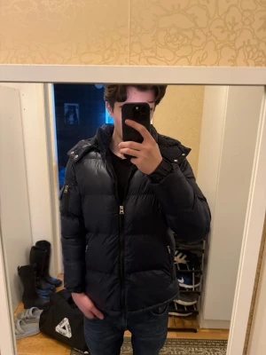 Mörkblå pufferjacka Brave Soul S - Säljer en mörkblå pufferjacka från Brave Soul i storlek S. Jackan har huva med tryckknappar, dragkedja framtill och på fickor, samt quiltad design. Materialet är glansig polyester och jackan har en normal passform. Perfekt för kalla dagar.