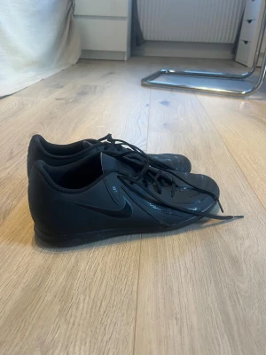 Nike Phantom inne fotbollsskor svarta - Nike Phantom inne fotbollsskor i helsvart med diskret Nike-logga på sidan och Phantom-tryck på hälen. Skorna har snörning och en rund tå, tillverkade i syntetmaterial med mönstrad ovansida för bättre bollkontroll. Perfekta för dig som vill ha stilrena och funktionella skor på planen. Strl 43 innermått 27,5 cm.