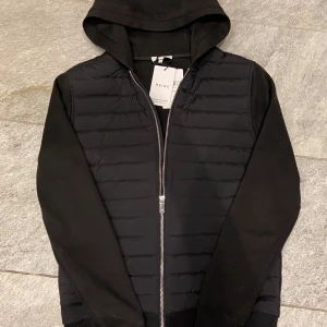 Svart cardigan från Reiss - Säljer en svart hybridjacka från Reiss i storlek XS men passar även S. Jackan har quiltad front, mjuka ärmar och huva. Den är typ helt ny och lapparna sitter kvar, nypris ligger på cirka 3 tusen o denna är som sagt typ helt ny så verkligen ett kap!👍