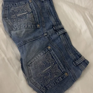 House of Denim jeansshorts med brodyr - Snygga blå jeansshorts från House of Denim med råa kanter och stora bakfickor dekorerade med broderi och blingdetaljer. Klassisk femficksmodell med knapp och dragkedja framtill. Perfekta för en chill sommarlook.