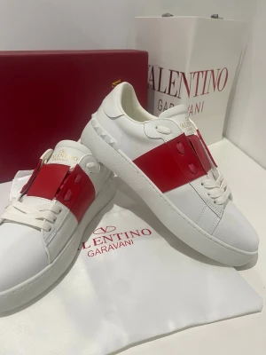 Valentino Garavani Open sneakers vit/röd - Snygga Valentino Garavani Open sneakers i vitt skinn med bred röd läderdetalj över mitten. Klassisk rund tå, platt sula och vita snören. Guldlogga på plösen och coola nitar baktill. Perfekta för dig som vill sticka ut med lyxig streetstyle.