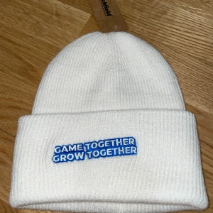 Vit ribbad mössa från Beechfield - Vit mössa från Beechfield med uppvikt kant och blå broderad text 'GAME TOGETHER GROW TOGETHER' framtill. Perfekt för kalla dagar och enkel att matcha med olika outfits. Presentkort ingår vid köp, Värde 59kr!