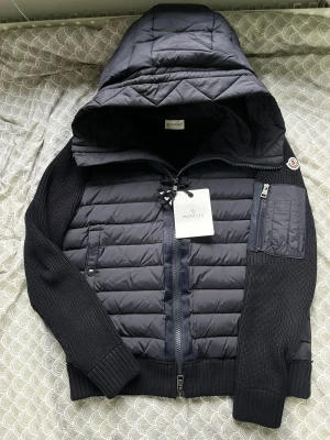  Moncler cardigan - Snygg Moncler med stickade ärmar och quiltad front. Jackan har en stor huva, dragkedja framtill, fickor med tryckknapp och en extra ficka med dragkedja på ärmen. Perfekt för dig som vill ha en stilren och exklusiv look.