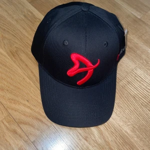 Svart keps från Arozzi med röd logga - Svart keps från Arozzi med böjd skärm och justerbart spänne bak. Framtill sitter en stor röd Arozzi-logga och på sidan finns Arozzi-texten i grått och rött. Presentkort ingår vid köp, värde 59kr!