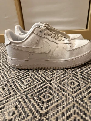 Nocta Air Force 1  - Säljer ett par snygga Nike Air Force 1 Low Drake NOCTA Certified Lover Boy, storlek 41, i helvitt skinn med snygga detaljer som broderad text på sulan och diskreta symboler på hälen. Klassisk siluett med snörning, perforerad tå och platt sula. Perfekta för dig som gillar stilrena sneakers med unik touch.