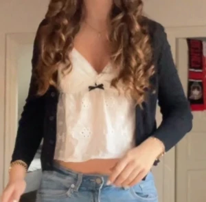 Vit broderad linnetopp med rosett - Supersöt vit linnetopp med broderade blommor och spetskant vid v-ringningen. Toppen har smala axelband och en liten svart rosett framtill som detalj. Croppad passform som ger en fräsch och somrig vibe. Perfekt att styla med jeans eller kjol!