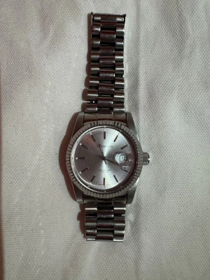 Silvrig klocka från Moretime - Stilren klocka från Moretime i helsilver med rund urtavla och datumvisning. Armbandet är i metall med klassisk länkdesign och spänne. Urtavlan är enkel och elegant med tydliga markeringar och diskret logga. Vattentålig upp till 50 meter. Saknar batteri. Extra delad kommer med 