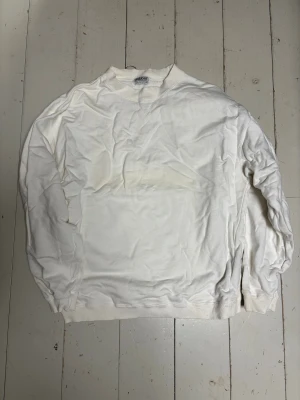 Vintage vit sweatshirt från Dobber - Vintage vit sweatshirt från Dobber med klassisk rund hals och ribbade muddar vid ärmslut och nederkant. Tröjan har en enkel och clean look, perfekt för dig som gillar basic och stilrent. Materialet är mjuk bomull och passformen är normal med långa ärmar. Den har en patch på ena ärmen som man enkelt kan rycka bort ifall min inte skulle vilja ha den. 