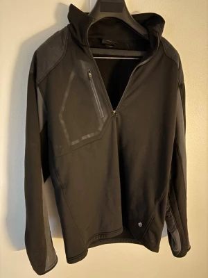 Svart vindjacka (golfjacka) från Galvin Green - Svart vindjacka från Galvin Green med hög krage och halv dragkedja. Jackan har en bröstficka med dragkedja och detaljer i mörkgrått på axlar och ärmar. Materialet är syntet och polyester, perfekt för blåsiga dagar. Snygg och sportig design med logga på kragen. Storlek: L passar M. Nypris: 2500