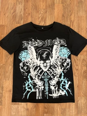 Svart t-shirt med grafiskt tryck XL - Svart t-shirt i storlek XL men sitter mer som ett M, med ett stort grafiskt tryck framtill i vitt och ljusblått. Trycket har dödskalle, vingar, rosor och texten 'DOOMED' i gotisk stil. Klassisk passform och rund halsringning.