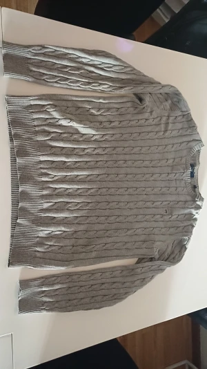 Grå kabelstickad tröja från GANT - Snygg grå kabelstickad tröja från GANT i premium bomull. Tröjan har rund hals, ribbade muddar och ett broderat GANT-emblem på bröstet. Perfekt för dig som gillar klassisk stil med en modern touch. Den är liten i storlek så skulle nogg säga att det är M men de beror på din längd. På ena hörnet på gant tagen i nacken så har det lossnat lite men inget man märker av, skriv för frågor o bilder.